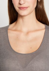 Närbild av en kvinnas nedre ansikte och nacke som bär en grå top med rundad halsringning och små ringörhängen.