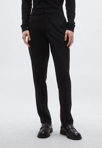 Čierne nohavice na mieru s úzkym strihom, vyrobené z hladkej látky, so zipsom na zapínanie a bočnými vreckami. Nosené s čiernymi nazúvacími topánkami.