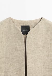 Beżowa tkanina lniana z dekoltem w serek i czarną metką z napisem „Massimo Dutti” wewnątrz kołnierza, wyprodukowana w Portugalii.
