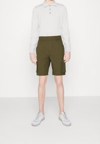 Shorts cargo kaki avec une taille élastique et deux poches latérales ; associés à un polo à manches longues gris clair et des baskets grises.