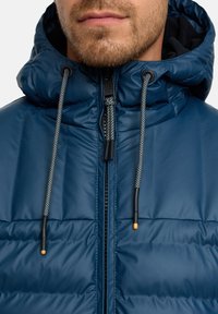 Mørkeblå polstret jakke med lynlås, hætte med snøre og grå accent-snøre. Har en glat, skinnende overflade og robust hardware.