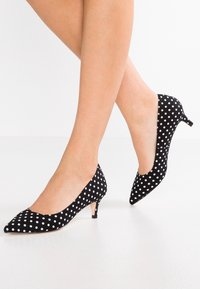Legs wearing black kitten heel shoes with white polka dots, standing against a plain white background.