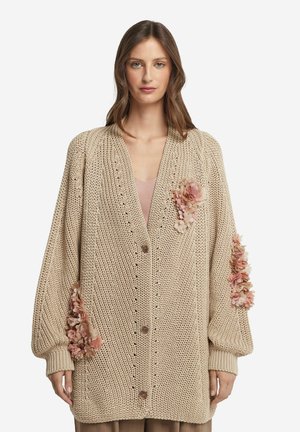 Beige Strickjacke mit lockerer Passform, großen Ärmeln, vorderen Knöpfen und stofflichen Blumenakzenten an beiden Ärmeln.