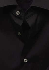 Camisa negra con un cuello estructurado, dos botones plateados y una textura de tela suave. El fondo es oscuro, realzando el color de la camisa.