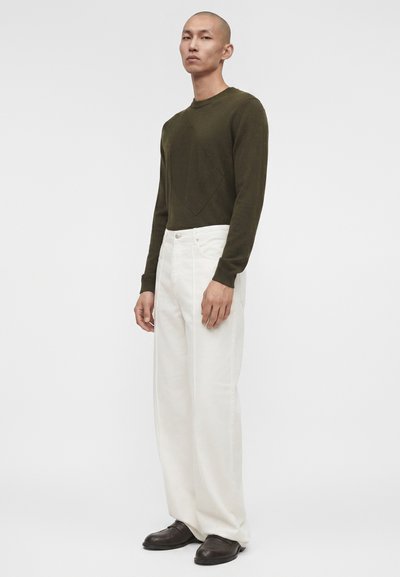 Άνδρας στέκεται φορώντας μια ελιάς μακρυμάνικη μπλούζα, λευκό παντελόνι με φαρδιές πόδιες και σκούρα καφέ παπούτσια σε λευκό φόντο.