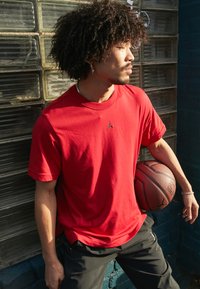 Camiseta vermelha de algodão com um pequeno logótipo preto, combinada com calças pretas. A pessoa segura uma bola de basquete contra uma parede texturizada.