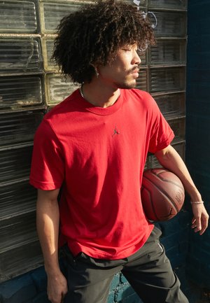 Rød bomuld t-shirt med et lille sort logo, parret med sorte bukser. Personen holder en basketball mod en tekstureret væg.