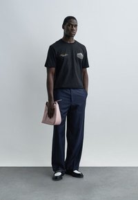 Νεαρός άνδρας που φοράει μαύρο γραφικό μπλουζάκι, μπλε παντελόνι με ρίγες, μαύρα και άσπρα μοκασίνια, κρατώντας μια ροζ τσάντα σε απλό φόντο.