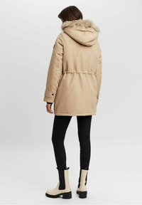 Parka beige con coulisse in vita e cappuccio con pelliccia sintetica, abbinata a leggings neri e stivaletti beige con suola chunky.