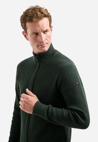 Bosgroene ritstruien met een textuurlijke breipatroon, hoge kraag en geribde manchetten. Bevat een klein hexagonaal logo op de schouder.