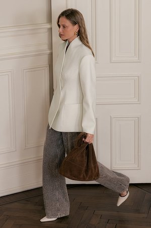 Witte op maat gemaakte blazer, losse grijze textuurbroek en een bruine suède tas, met puntige beige schoenen, voor een neutrale muur.