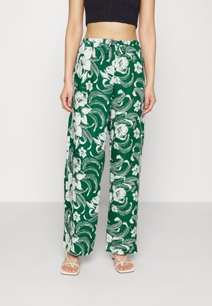 Vrouw draagt groene wijde broek met witte bloemprint, een zwart cropped topje en witte sandalen met bandjes, staand tegen een effen muur.