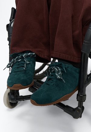 Personne portant des baskets vert foncé et un pantalon marron assise dans un fauteuil roulant noir montrant les repose-pieds et les roues.