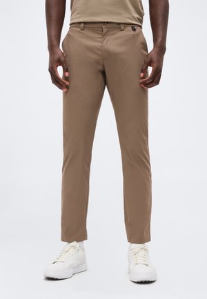 Homme portant un pantalon slim beige, des chaussettes blanches et des baskets blanches, debout les mains détendues le long du corps sur un fond uni clair.