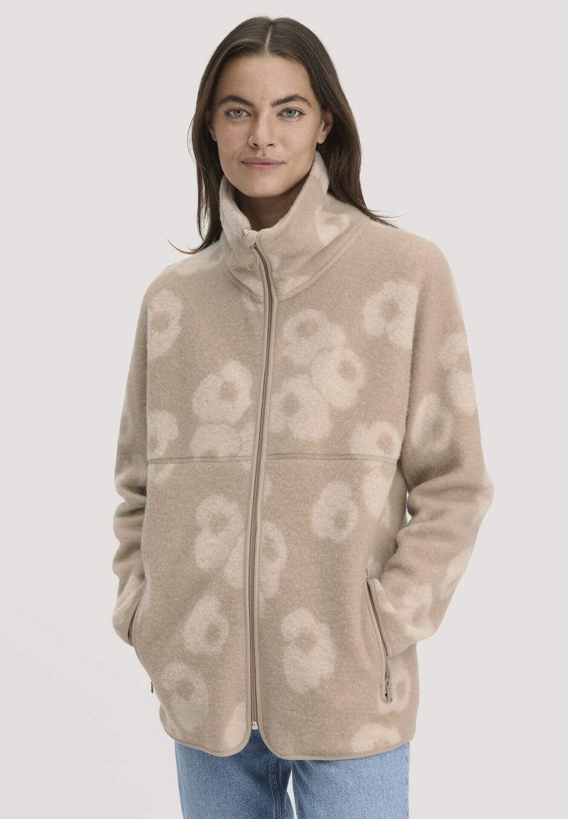 Vrouw in een beige fleecejack met ritssluiting en hoge kraag, met een licht bloemenpatroon, handen in de zakken, gecombineerd met blauwe denim.