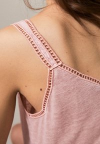 Jasnoróżowy top na ramiączkach z delikatnym koronkowym wykończeniem przy ramiączkach, wykonany z miękkiego materiału o luźnym kroju.