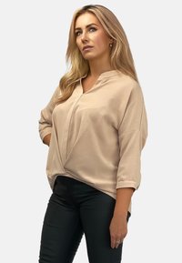 Beżowa bluzka z dekoltem w serek i rękawami o długości 3/4, z subtelnym połyskiem i drapowaniem. W połączeniu z czarnymi dopasowanymi spodniami.