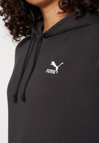 Femme portant un sweat à capuche noir Puma avec cordons de serrage et logo Puma blanc sur la poitrine, montrant ses cheveux bouclés et son cou.