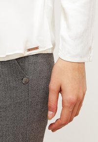 Weiße Bluse mit langen Ärmeln, abgerundetem Saum und kleinem Metall-Logoanhänger; kombiniert mit grauen Fischgrat-Hosen, die einen glänzenden Knopf haben.