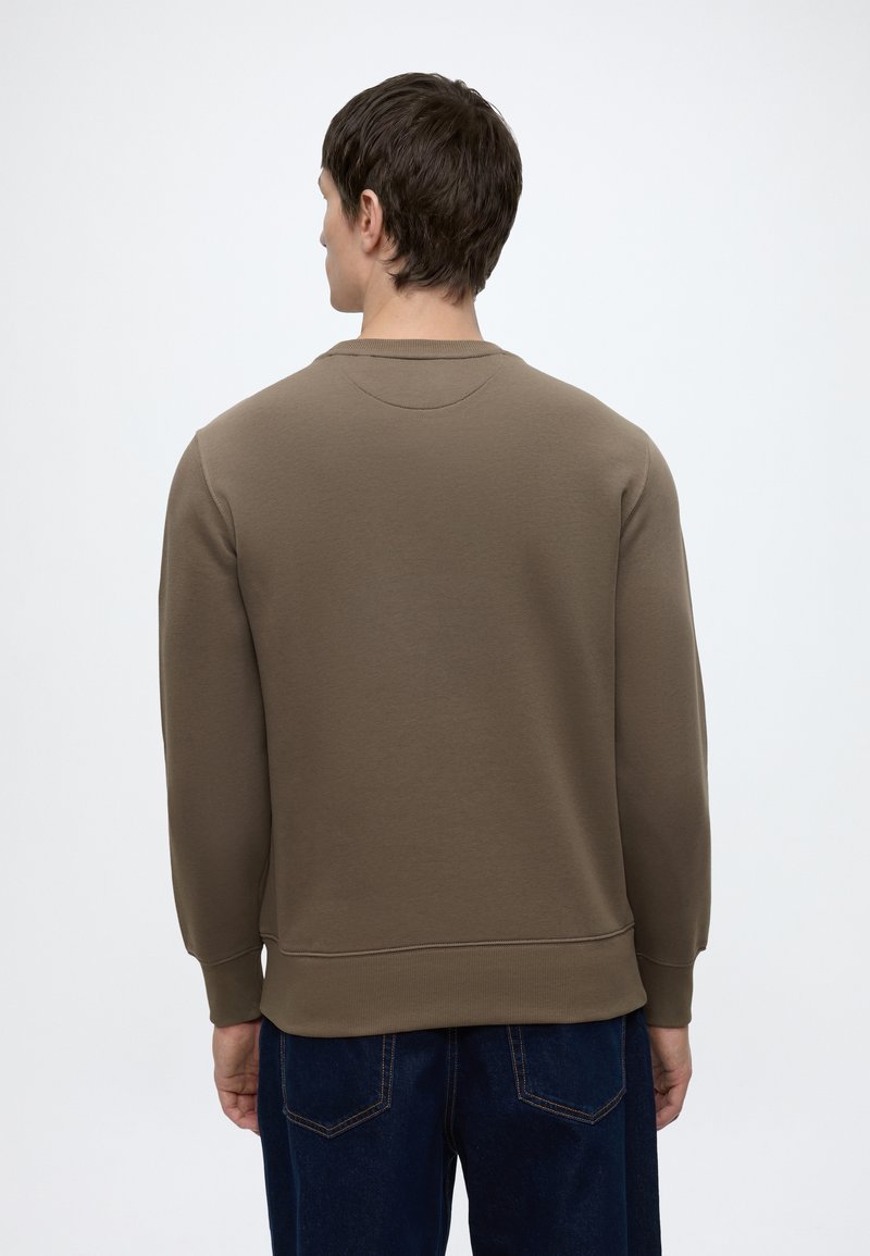 Mand set bagfra iført en ensfarvet brun langærmet sweatshirt og mørkeblå jeans mod en hvid baggrund.