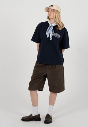 Vrouw met beige pet, navy T-shirt, blauw patroonige halsdoek, bruine knielange shorts, witte sokken en bruine loafers poseert tegen een witte achtergrond.