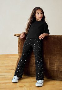 Mustast lõikega pluus, mille juurde on kombineeritud lehvipükstega mustad püksid, millel on valge tähe muster, täiendatud valgete tossudega. Taustal tekstuuriga pruun diivan.