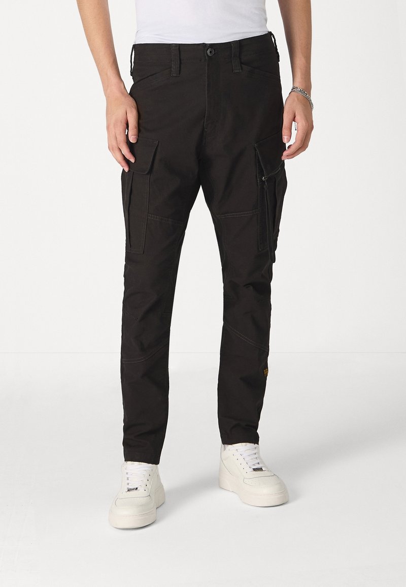 Pantalon cargo noir avec plusieurs poches porté avec des baskets blanches et une chemise blanche, vue de face de la taille aux pieds.