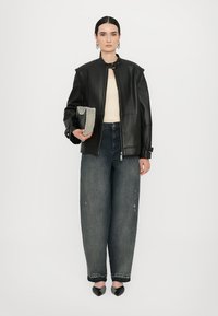 Giacca in pelle nera con colletto alto, top crema, jeans blu oversize e scarpe nere; tiene una borsa a tracolla con borchie.