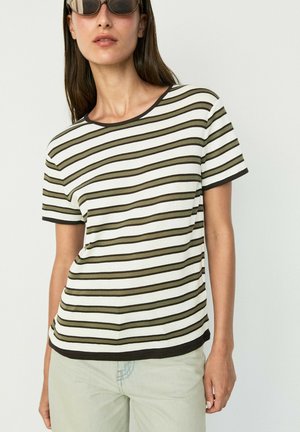 Frau mit Sonnenbrille, einem weißen T-Shirt mit kurzen Ärmeln und schwarzen sowie olivgrünen horizontalen Streifen und hellen Hosen.