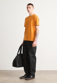 Orange bomulds t-shirt med korte ærmer, parret med mørke denim bukser og sorte sneakers. Holder en sort taske med skulderstropper.
