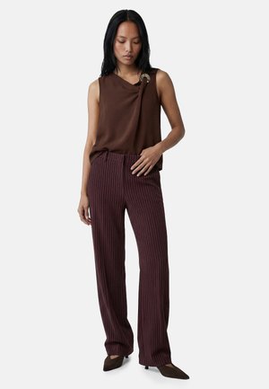 Frau trägt ärmelloses braunes Top mit Ringdetail und hoch taillierte, weit geschnittene braune Nadelstreifenhose, steht mit einer Hand in der Hüfte.