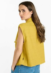 Un haut sans manches texturé jaune avec un motif en relief subtil, doté d'un col montant et de poches latérales, associé à un bas en denim.