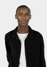 Svart, zip-up jakke med krage-design og brystlomme, båret over en hvit t-skjorte med rund hals. Glatt tekstur med minimal detaljering.