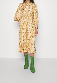 Abito midi a stampa floreale in crema e oro, con maniche a sbuffo e scollatura alta. Abbinato a stivali al ginocchio in suede verde.