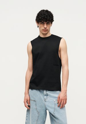 Mladý muž s kučeravými vlasmi, oblečený v čiernom tielku bez rukávov a svetlomodrých voľných džínsoch, stojaci pred jednoduchým svetlým pozadím.