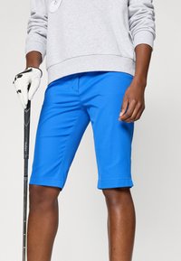 Blå golfshorts laget av lett stoff, med en slim fit og rullede ermer. Kombinert med en grå hettegenser og golfklubbe.