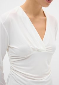 Blusa bianca a maniche lunghe in tessuto leggero, caratterizzata da una scollatura a V e dettagli arricciati sui lati per una silhouette aderente. Texture liscia.