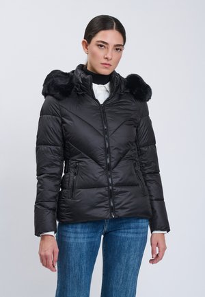 Frau trägt eine schwarze gesteppte Jacke mit pelzbesetzter Kapuze, weißes Hemd und blaue Jeans, steht vor einem einfarbigen hellen Hintergrund.
