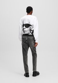 White sweatshirt featuring a black graphic of a man in sunglasses on the back. Worn with gray denim jeans and black shoes.