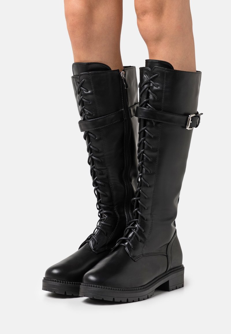 Bottes hautes en cuir noir avec lacets à l'avant, fermeture éclair latérale, sangle à boucle réglable et semelle épaisse portées sur des jambes nues.