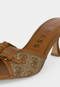 Mule à talon haut en cuir marron et tissu à motifs avec une boucle dorée et le nom de la marque "GUESS" sur la semelle intérieure.