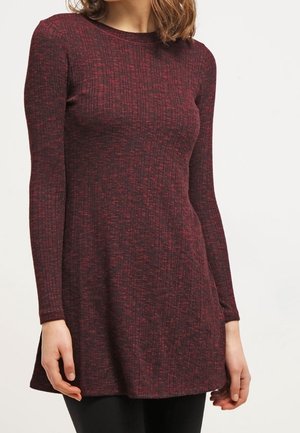 Femme portant un pull tunique ajusté à manches longues en maille côtelée rouge foncé et noir, debout devant un fond uni.