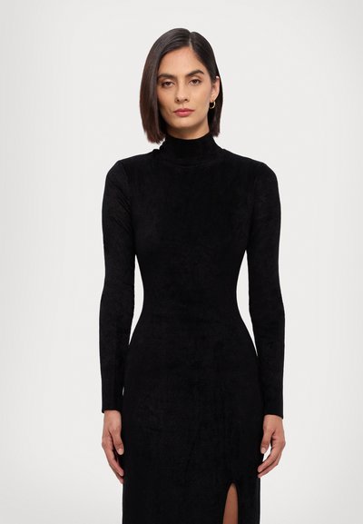 Robe noire à manches longues avec un col haut, une silhouette ajustée, une texture douce et des fentes sur les côtés.
