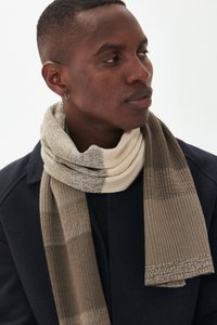 Homme portant une écharpe en tricot beige et grise enroulée autour du cou, vêtu d'un manteau sombre, regardant sur le côté sur un fond blanc.