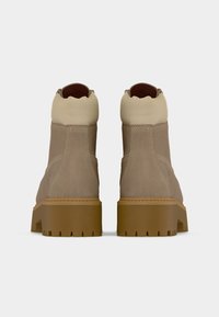 Ζευγάρι από μποτάκια αστραγάλου σε μπεζ και καφέ σουέντ με χοντρές λαστιχένιες σόλες, εμφανιζόμενα από πίσω σε λευκό απλό φόντο.