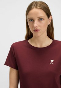 Burgundy t-shirt made of cotton, featuring a round neckline and short sleeves. Front has a white heart and text accent. Smooth texture.