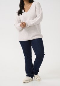 Lys pink sweater med V-hals, der har subtile glimmerende elementer, parret med mørkeblå jeans og hvide sneakers.