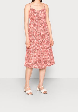 Frau trägt ein ärmelloses, rotes Midikleid mit weißem Punktmuster und Knöpfen vorne, kombiniert mit weißen Sandalen mit Absatz.