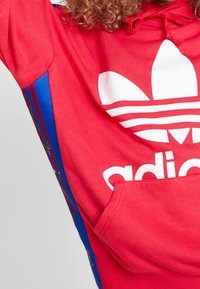 Röd hoodie med en stor vit Adidas trefoil-logga. Har blå sidostrippar med ett blommönster. Mjukt tyg och känguruficka.