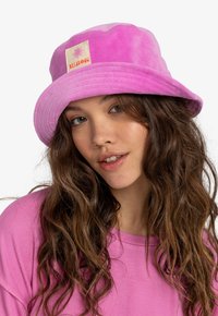 Cappello a secchiello rosa realizzato in tessuto morbido e con trama, con una toppa quadrata sul davanti che presenta un'illustrazione di sole e un bordo rigato.